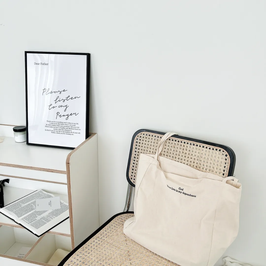  God Your love is my dependence canvas tote bag in cream, resting on a rattan chair beside a minimalist desk with an inspirational framed prayer print, creating a peaceful and uplifting space.