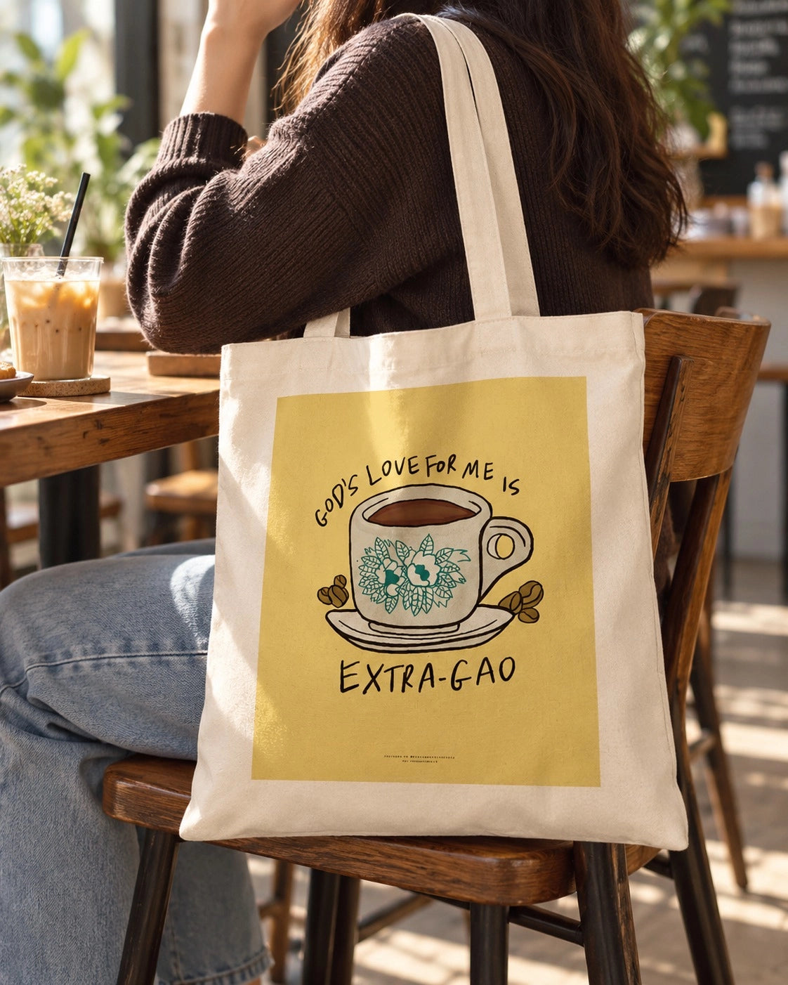 God's Love for Me Is Extra Gao cream tote bag with illustrated coffee cup graphic styled at a cosy cafe.