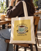 God's Love for Me Is Extra Gao cream tote bag with illustrated coffee cup graphic styled at a cosy cafe.