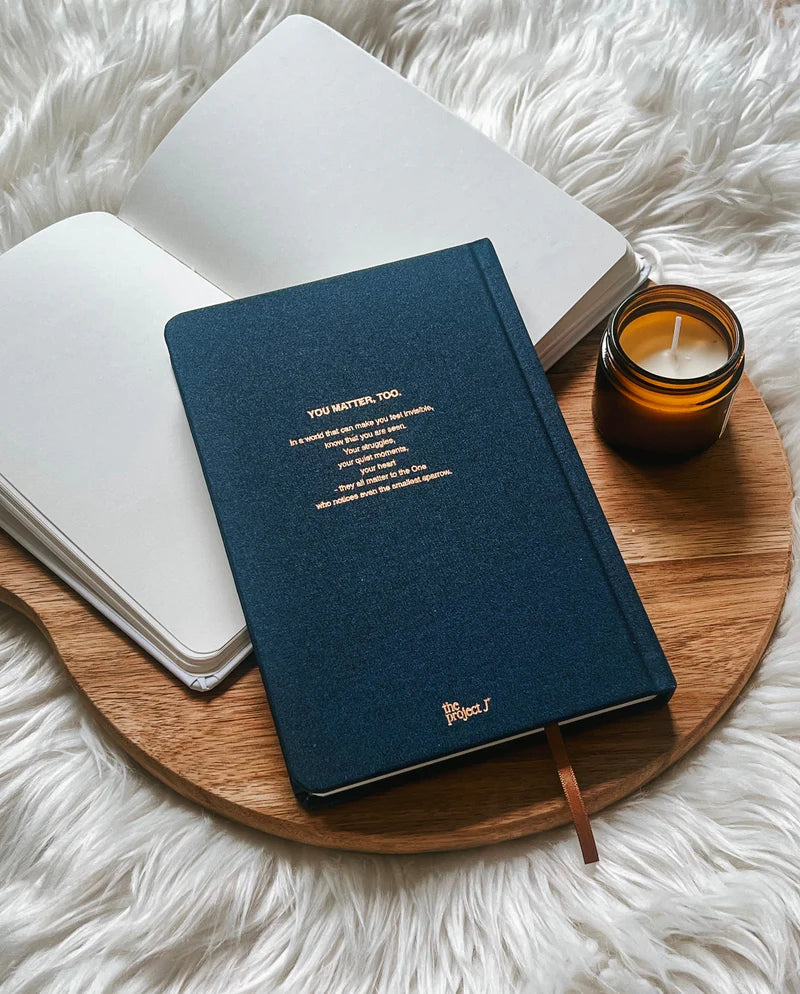 Blue fabric journal with gold text design on back cover by Project J placed on wooden tray beside open notebook and candle.