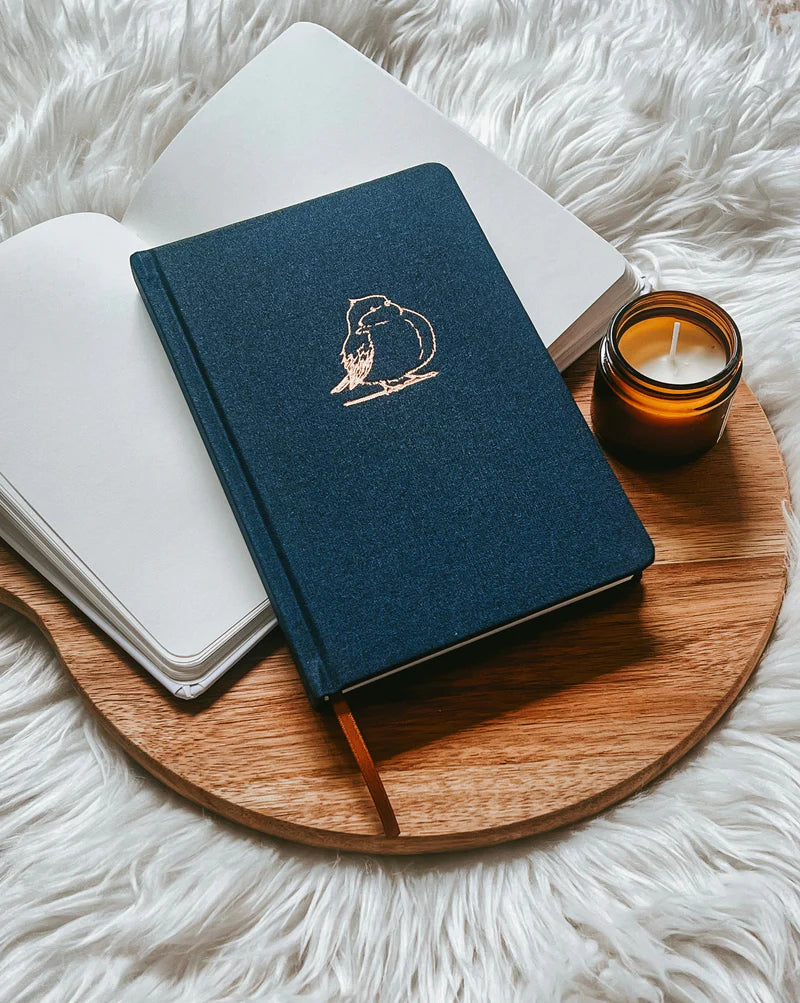 Blue fabric hardcover journal by Project J with gold bird design, placed on wooden tray beside open notebook and candle.