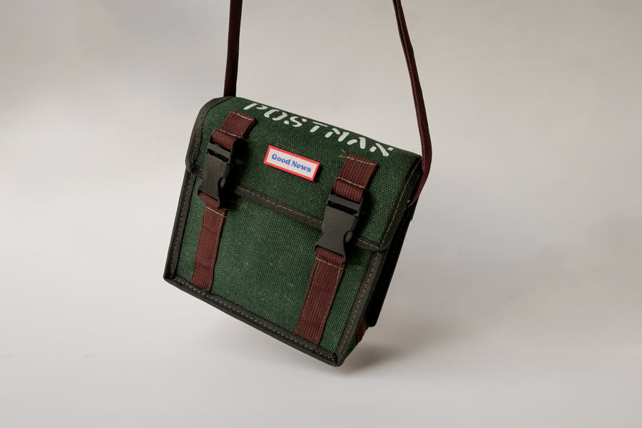 A green canvas messenger bag with 'Good News' branding, featuring brown straps and sturdy clips. The bag has a faith-inspired design and is ideal for daily use, with a minimalist style and practical functionality.