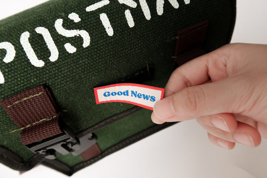 A close-up view of a green canvas messenger bag with the label 'Good News' being attached. The bag has 'POSTMAN' text on the top and a faith-based theme, showcasing both practicality and inspiration.