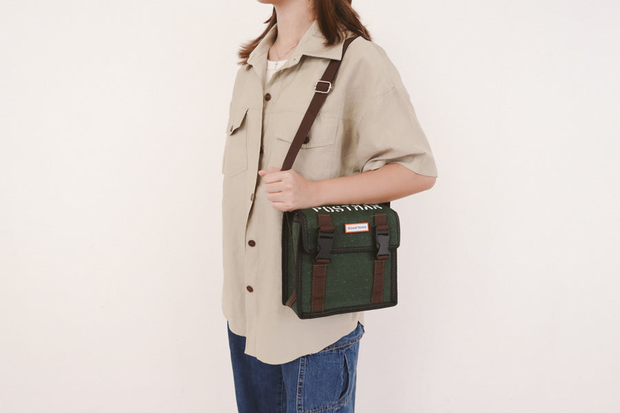 A person wearing a green canvas messenger bag with 'Good News' branding, shown from the side with the bag worn crossbody. The bag features brown straps and clips, offering a practical and faith-inspired design.