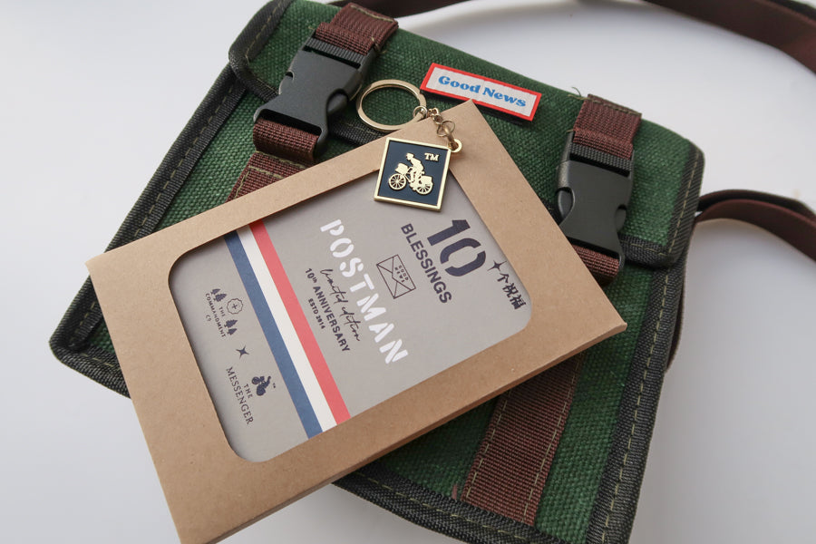 Close-up of a green canvas 'Good News' messenger bag with 'POSTMAN' text, displayed with a 10th anniversary edition 'Postman' keychain and packaging. The bag’s brown straps complement the faith-inspired design.