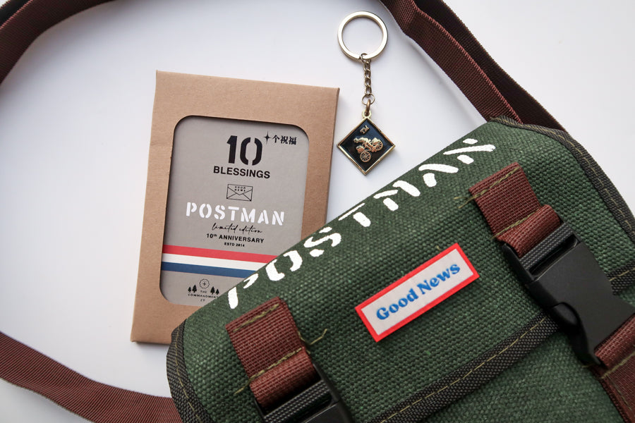 A green canvas 'Good News' messenger bag with brown straps displayed alongside the 10th anniversary edition 'Postman' keychain and packaging. The design features a bold 'POSTMAN' label on the bag and a small motorbike-themed keychain.