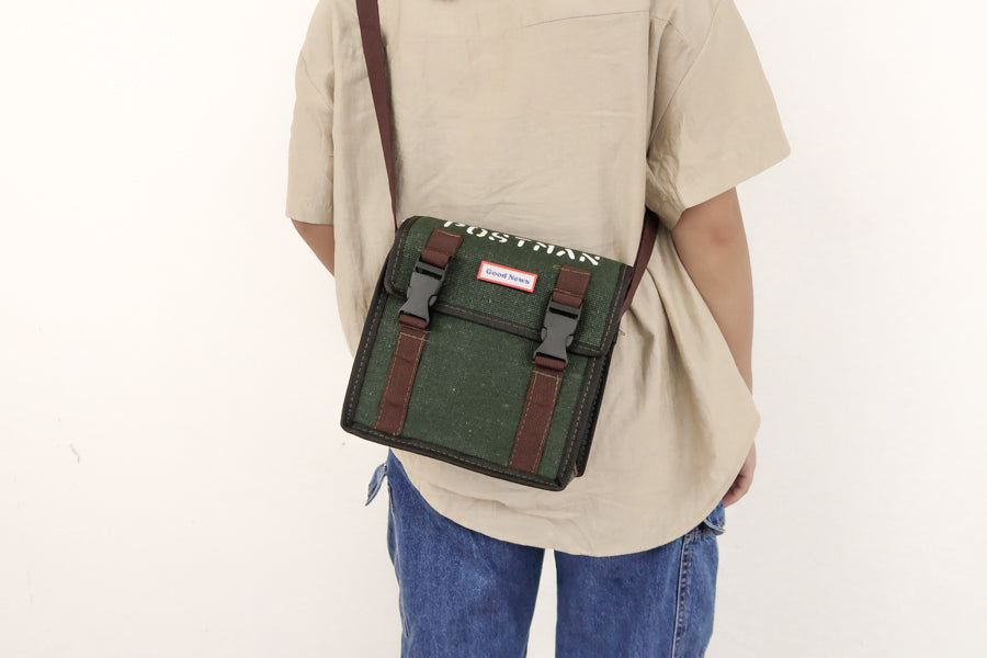 A rear view of a person wearing a green canvas messenger bag with 'Good News' branding. The bag is faith-themed with 'POSTMAN' text on top and brown straps, suitable for everyday use.