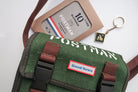 A close-up of a green canvas messenger bag with 'Good News' branding, featuring a 10th anniversary edition 'Postman' keychain and packaging. The brown straps and keychain complement the faith-inspired design.