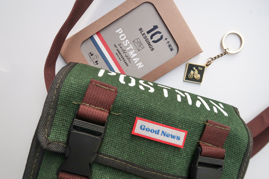 A close-up of a green canvas messenger bag with 'Good News' branding, featuring a 10th anniversary edition 'Postman' keychain and packaging. The brown straps and keychain complement the faith-inspired design.