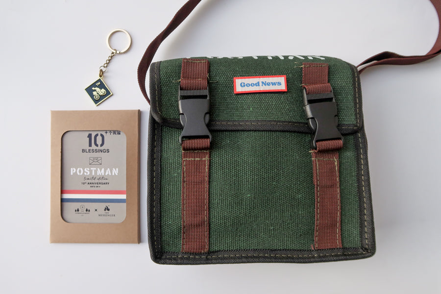 A green canvas messenger bag with 'Good News' branding displayed alongside the 10th anniversary 'Postman' keychain and packaging. The bag has brown straps and a faith-themed design, symbolizing both practicality and inspiration.