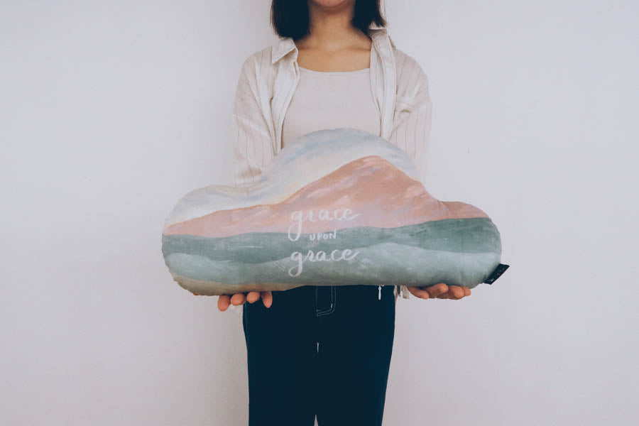 Person holding a cloud-shaped plush pillow with the phrase 'grace upon grace,' symbolizing faith and comfort in a soft decorative cushion.