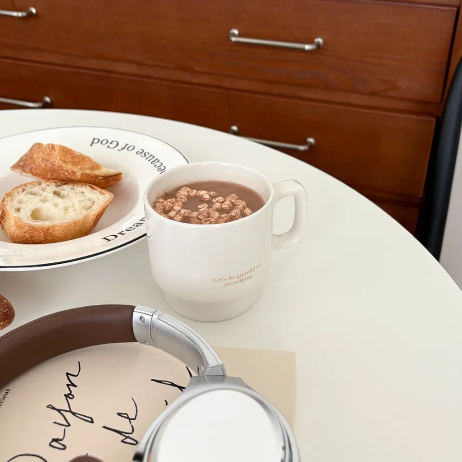 White mug with the words "Let's be grateful to everything" filled with hot chocolate and cereal, placed on a table with bread, a plate, and headphones, inspiring gratitude and simple happiness as a Christian gift from Jincha God.