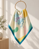 Cream Christian scarf with colourful Grace text and teal border draped over a wooden ring in a sunny room.