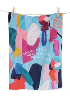 Colorful abstract art tea towel with pastel blues, pinks, and orange shapes, featuring the curved white text “Greatly Blessed, Highly Favored, Deeply Loved” on a light blue background, hanging on a clothesline.
