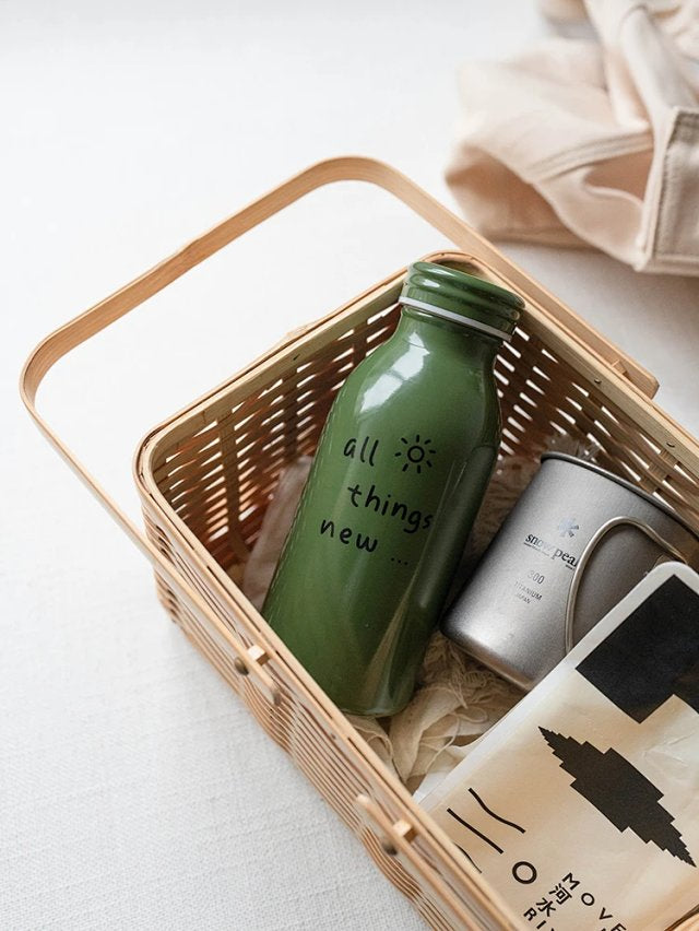 Green milk-bottle shaped stainless steel tumbler with the text “all things new,” placed in a woven basket with a mug and magazine—faith-inspired lifestyle gift by The Commandment Co.