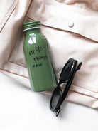 Green milk-bottle shaped stainless steel tumbler with the "all things new" message, placed in a lifestyle setting beside a beige bag and black sunglasses—faith-inspired, uplifting gift-ready imagery for The Commandment Co.