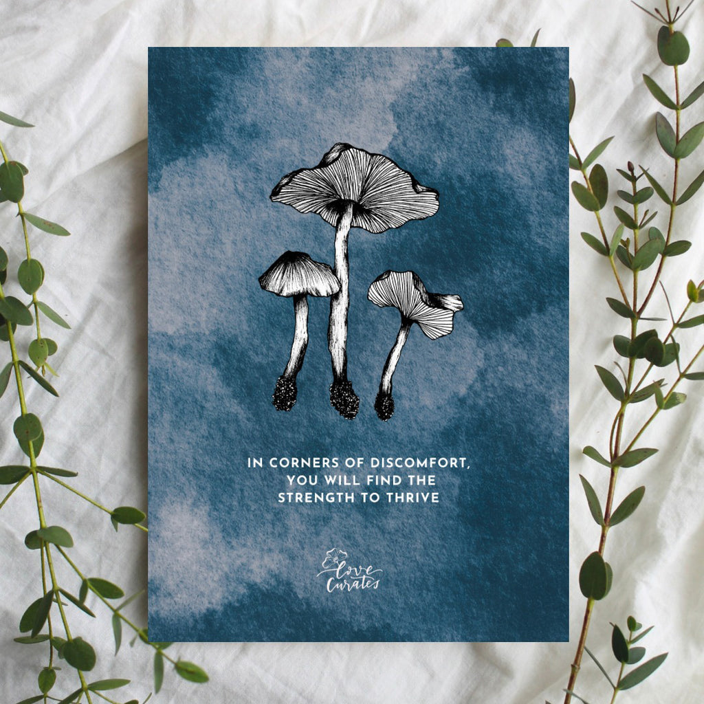 A greeting card featuring a black and white illustration of three mushrooms on a textured blue background. The card displays the quote 'In corners of discomfort, you will find the strength to thrive,' conveying a message of resilience and growth. Green foliage frames the card.