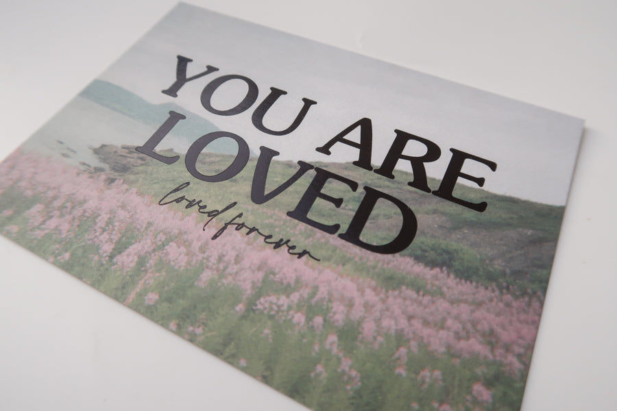 A detailed view of a postcard with a scenic field of pink flowers and the heartfelt message "You Are Loved, loved forever," offering reassurance and comfort.