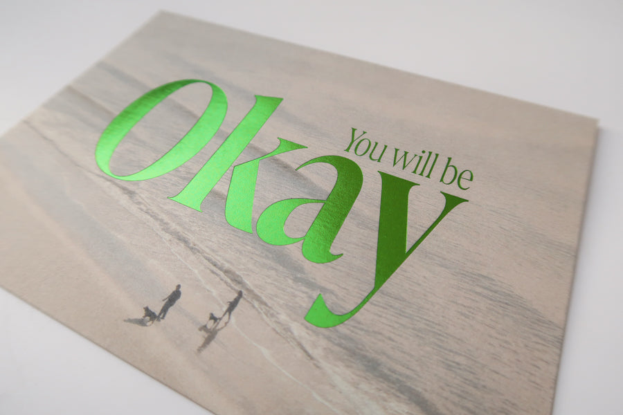 A detailed view of a postcard with a serene beach background and the comforting text "You Will Be Okay" in bold green lettering, designed to offer reassurance and encouragement.