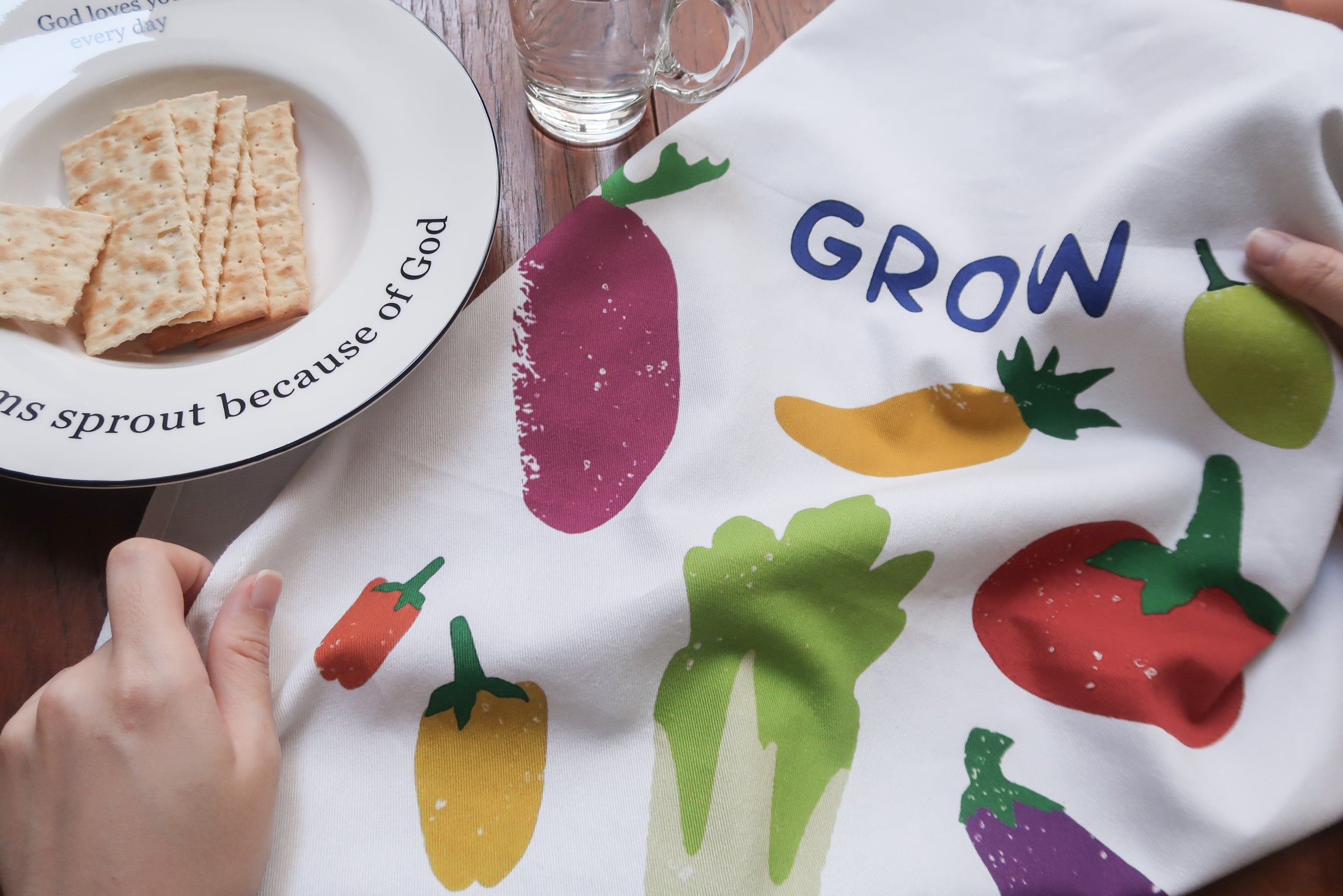 Grow in Grace tea towel with a colorful vegetable print on white fabric, shown beside a plate of crackers on a wooden table—a contemporary Christian kitchen decor gift.