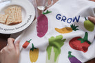 Grow in Grace tea towel with a colorful vegetable print on white fabric, shown beside a plate of crackers on a wooden table—a contemporary Christian kitchen decor gift.