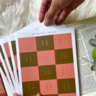 Hand holding sheets of pink and gold Bible tabs labeled with books like Genesis and Joshua, alongside an open Bible showing the book of Genesis with eucalyptus leaves on a soft white background.