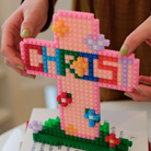 Hands holding a colorful micro-brick Christian cross with “CHRIST” lettering and flower accents, faith-inspired DIY decor from The Commandment Co