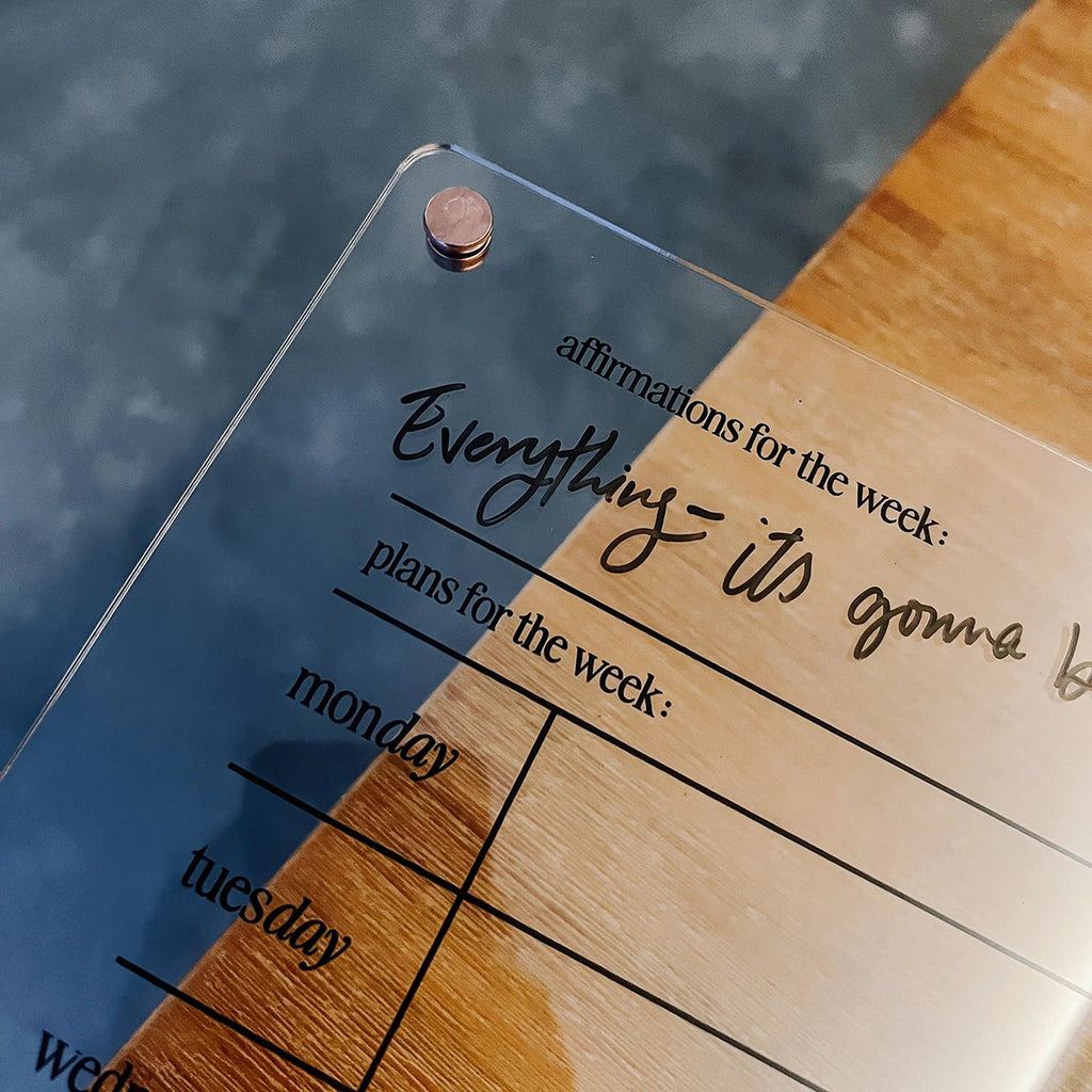 Handwritten affirmation 'Everything - it's gonna be...' on a transparent acrylic weekly planner board placed on a wooden surface.