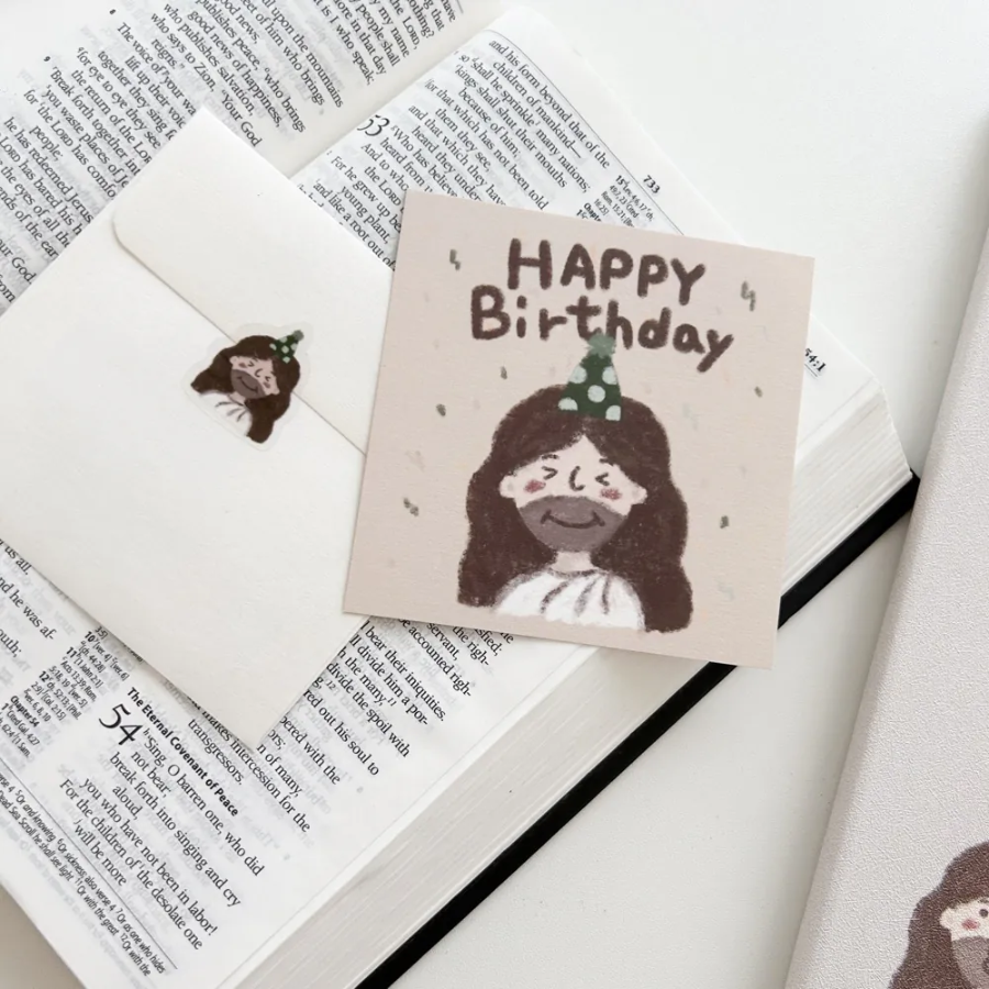 "Happy Birthday" postcard with a cute illustration of Jesus in a party hat, paired with a matching envelope and sticker, placed on an open Bible, sharing encouragement and joy.