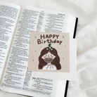 "Happy Birthday" postcard featuring a cute illustration of Jesus with a party hat, placed on an open Bible.