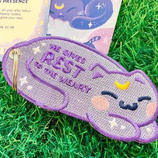 Purple embroidered cat keychain with the Bible verse 'He gives rest to the weary,' featuring a smiling cat design with blush cheeks, moon detail, and stars, displayed on green grass.