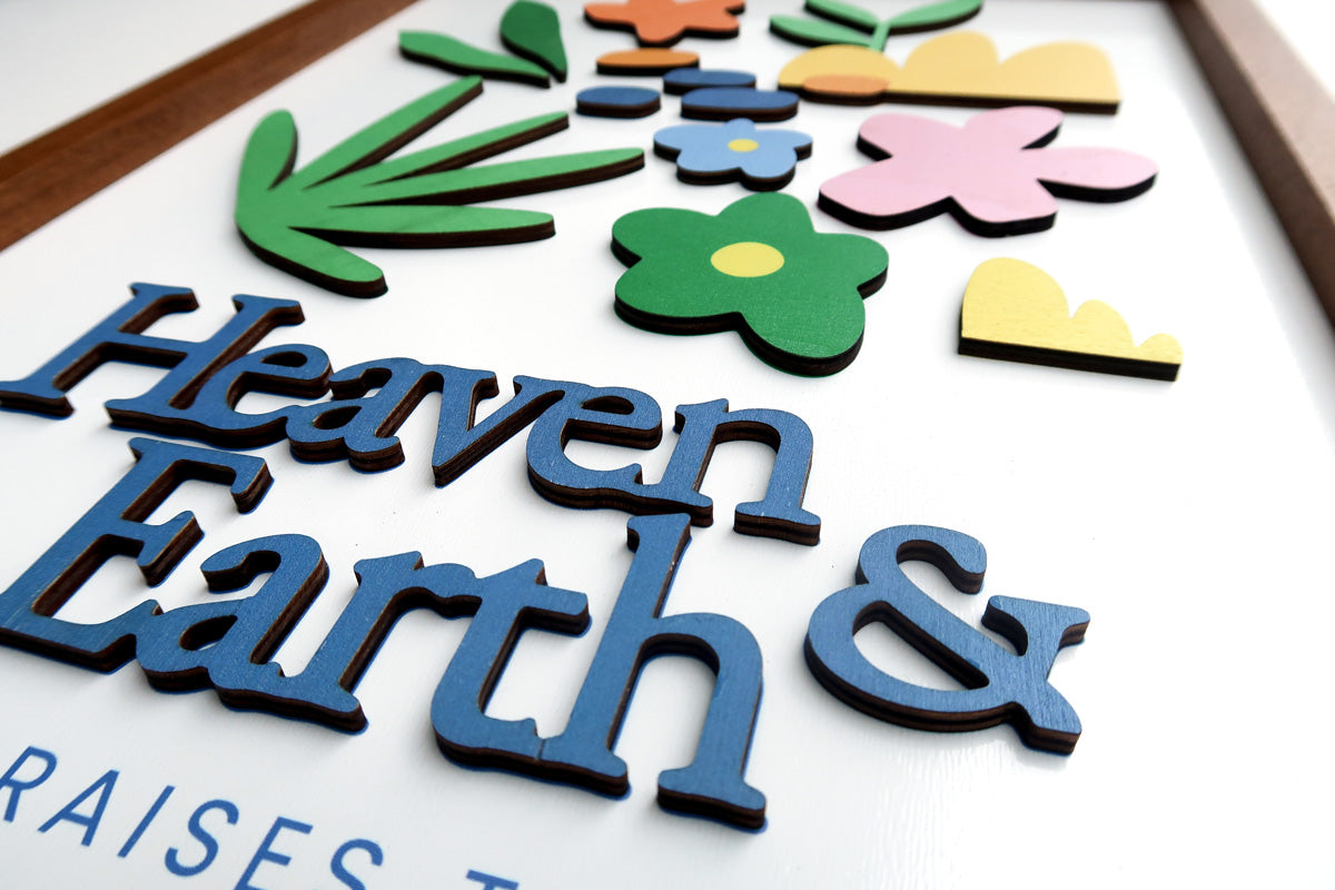 Close-up of a wooden scripture poster featuring the words "Heaven & Earth" with colorful floral and plant designs in a layered, handcrafted style, perfect for faith-inspired home decor.