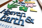 Close-up of a wooden scripture poster featuring the words "Heaven & Earth" with colorful floral and plant designs in a layered, handcrafted style, perfect for faith-inspired home decor.