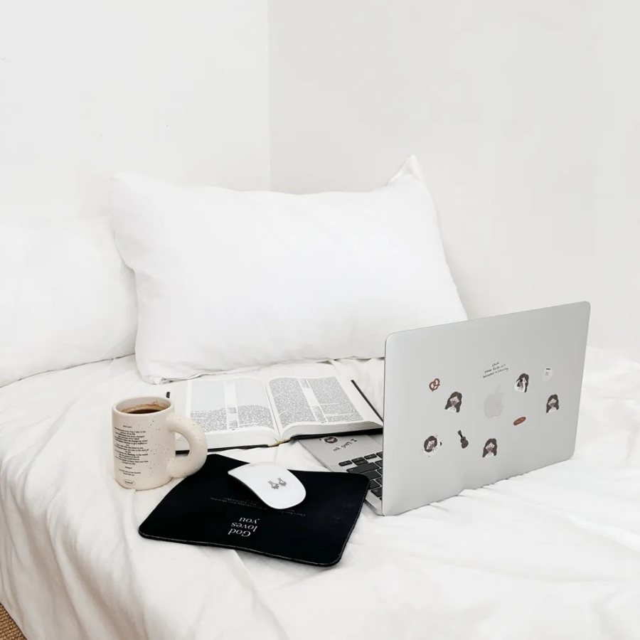 Cozy workspace on a bed with a laptop decorated with "Hey! I Love You" Jesus stickers, an open Bible, a coffee mug, and a "God loves you" mouse pad, inspiring encouragement and simple happiness.