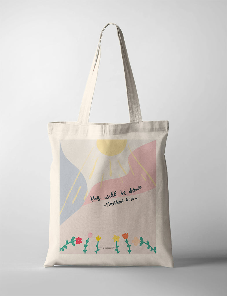 
A beige tote bag featuring the message "His will be done - Matthew 6:10," with a vibrant design of a sun, pastel hills, and colorful flowers, creating a cheerful and inspirational look.