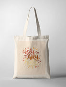 A faith-based Tote Bag featuring the words 'Holy, Holy, Holy' in warm, earthy typography with red and gold hand-drawn hearts and decorative dots. The minimalist Christian wall art has a soft beige background, framed in a simple wooden frame, making it a perfect decor piece for a faith-inspired home or prayer space.