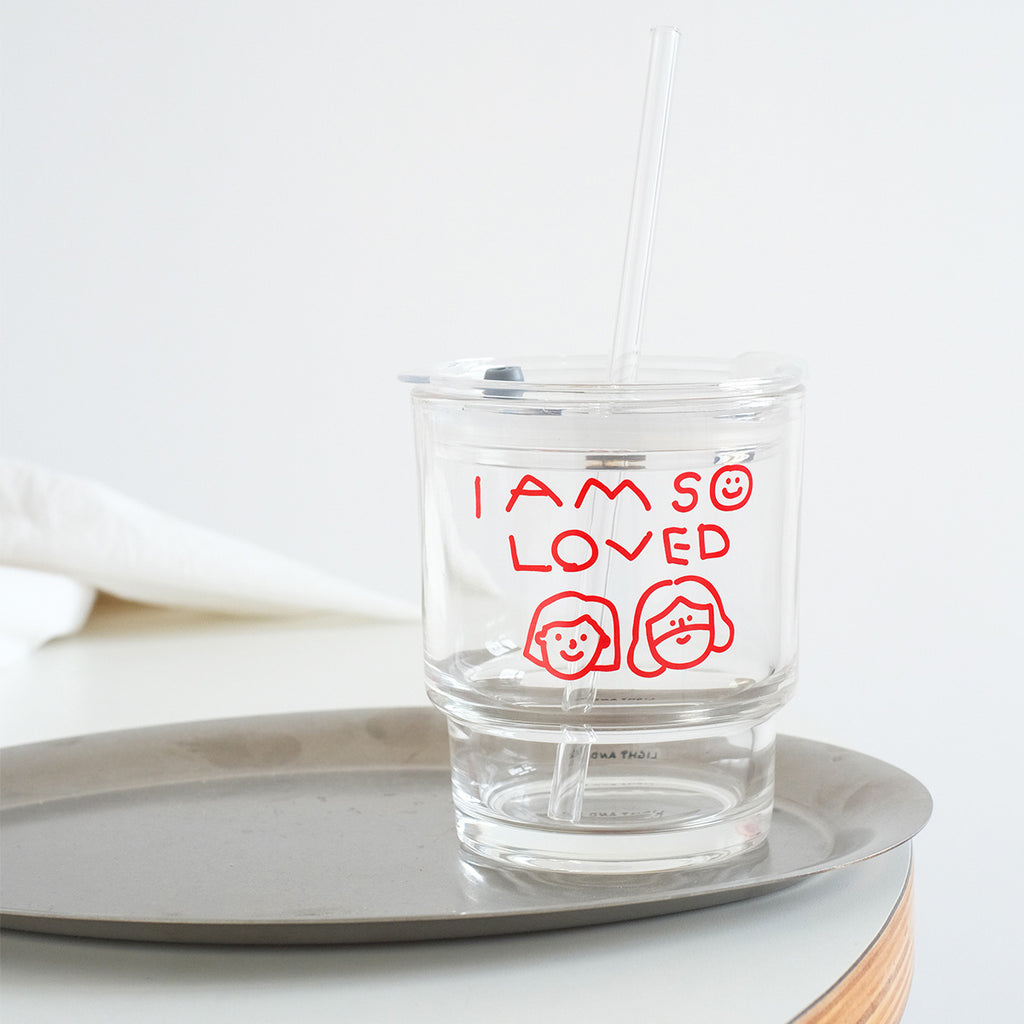 Clear glass tumbler with red text saying 'I Am So Loved' and hand-drawn smiley faces, featuring a reusable straw and eco-friendly minimalist design.