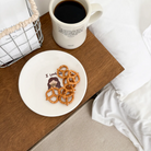 I Love You" dessert plate with a cute illustration of Jesus and pretzels, placed on a wooden bedside table next to a coffee mug, inspiring love and simple happiness as a Christian gift from Jincha God.