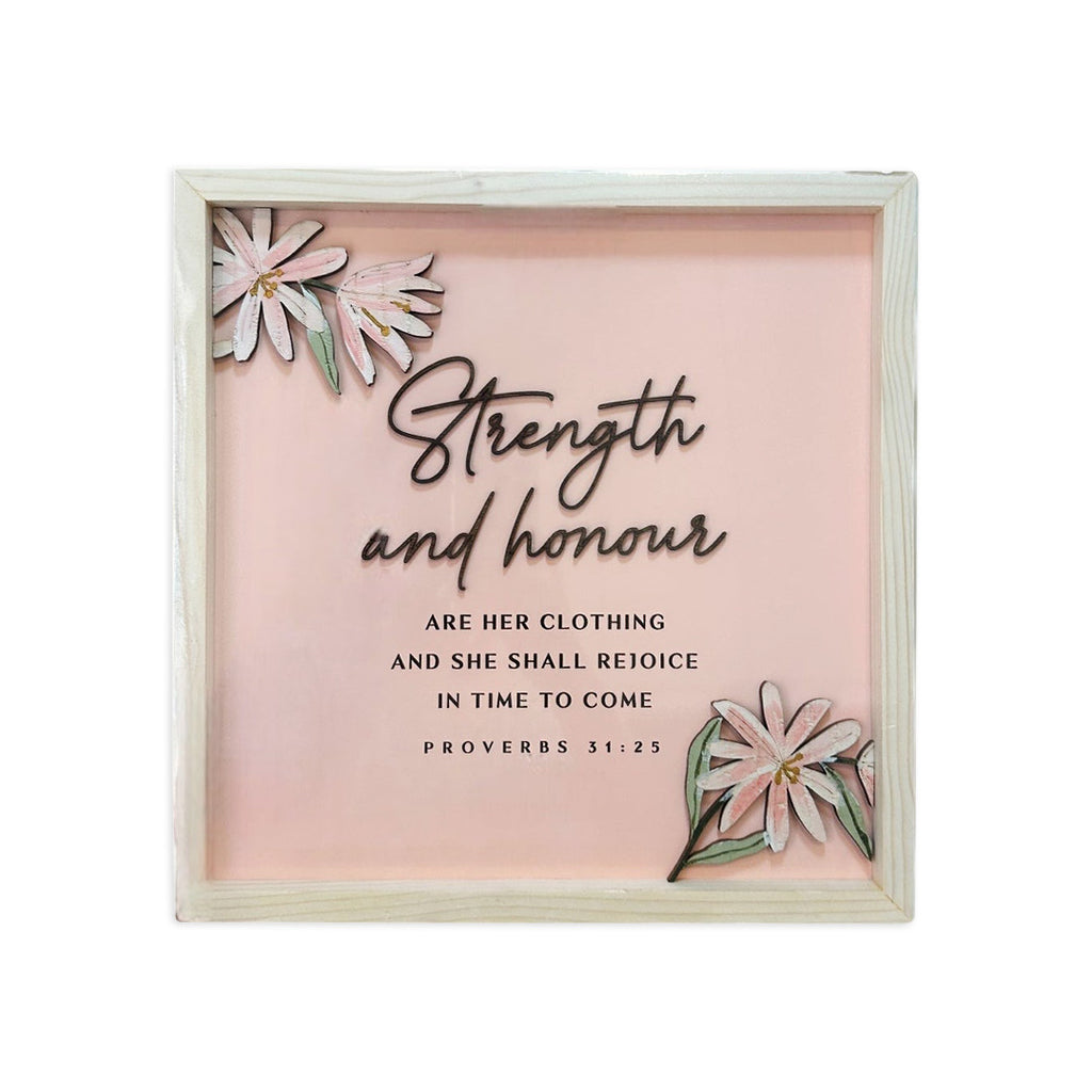 A framed Christian wall art featuring the Bible verse 'Strength and honour are her clothing and she shall rejoice in time to come' from Proverbs 31:25. The design is displayed on a soft pink background, with decorative floral elements in the corners, and is encased in a wooden frame.