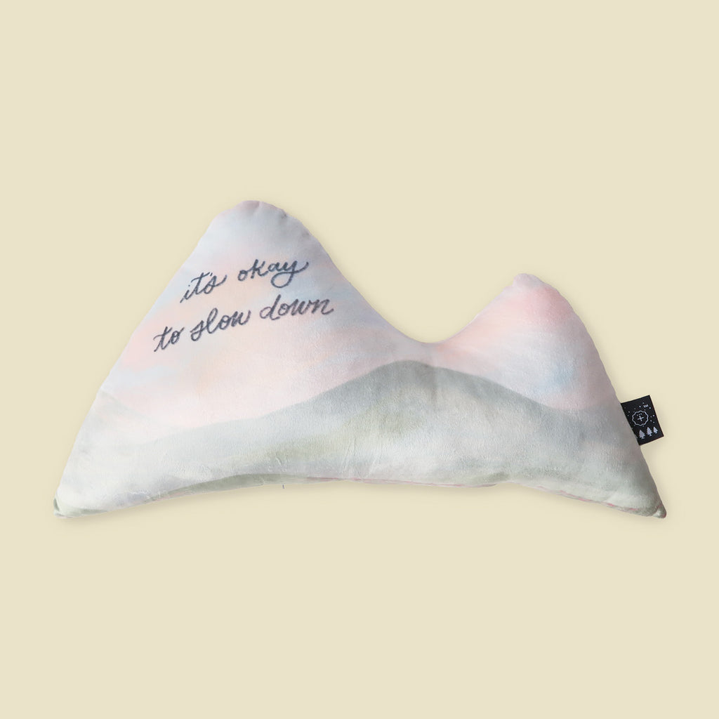 Pastel-colored plush pillow shaped like a mountain, featuring the uplifting message 'it's okay to slow down,' ideal for creating a peaceful, comforting space.