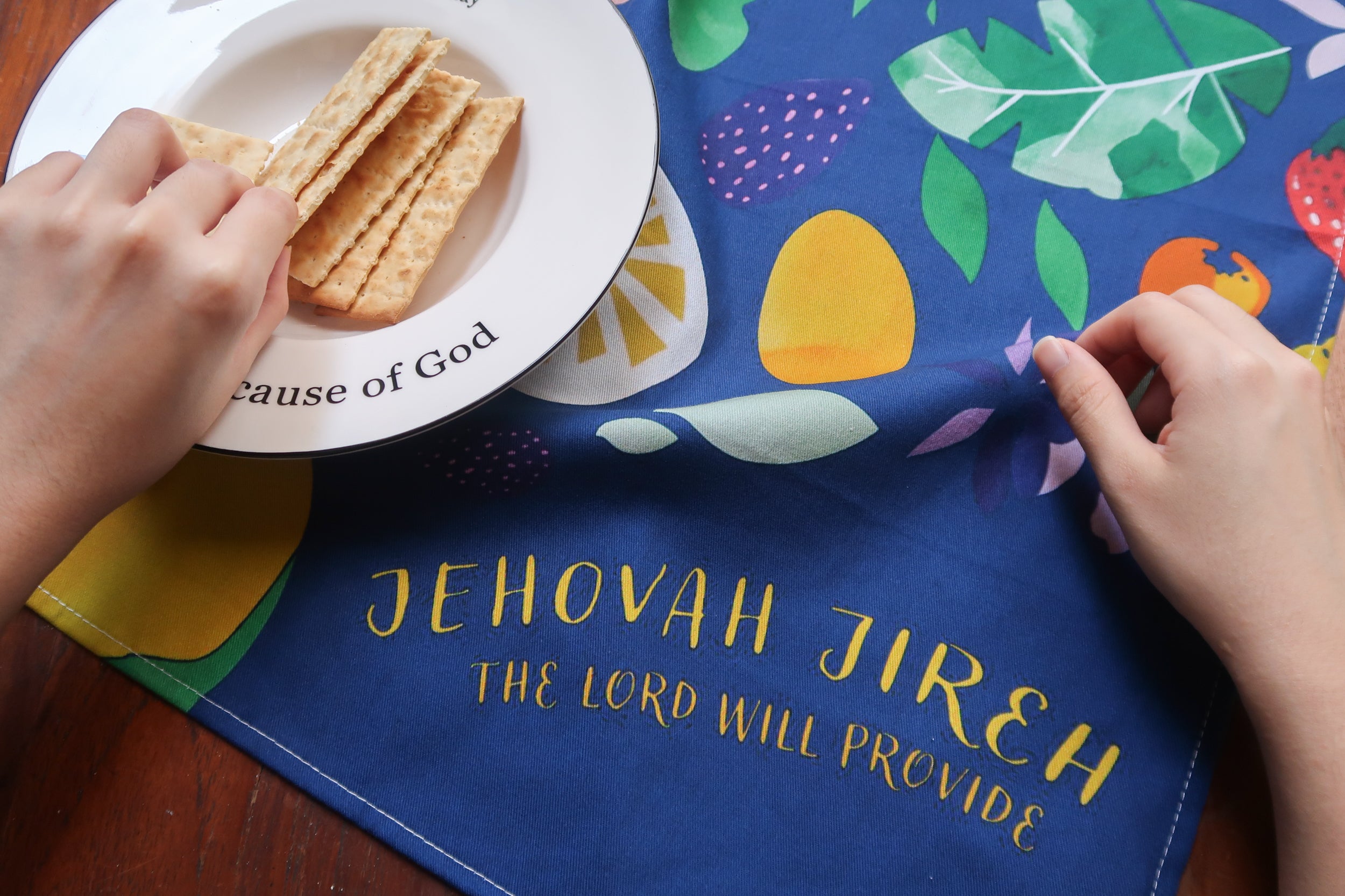 Jehovah Jireh tea towel with a blue background, colorful fruit pattern, and gold verse "The Lord Will Provide," shown with hands and a plate of crackers on a wooden table.