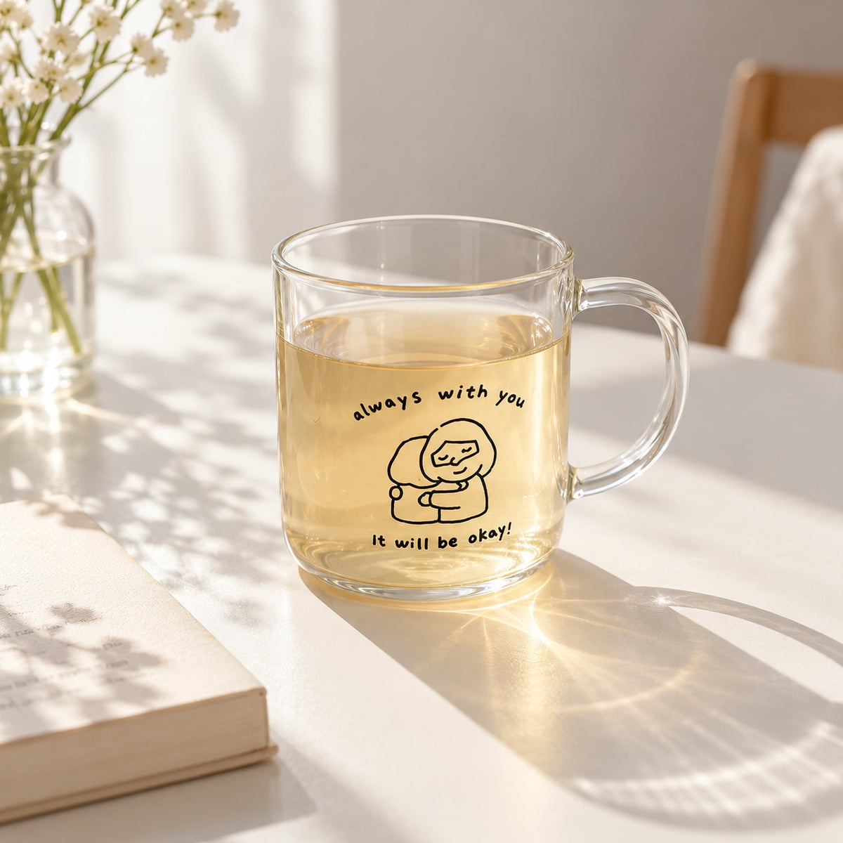 Clear glass mug with Always with you, it will be okay Christian illustration, filled with tea on a bright sunlit table.