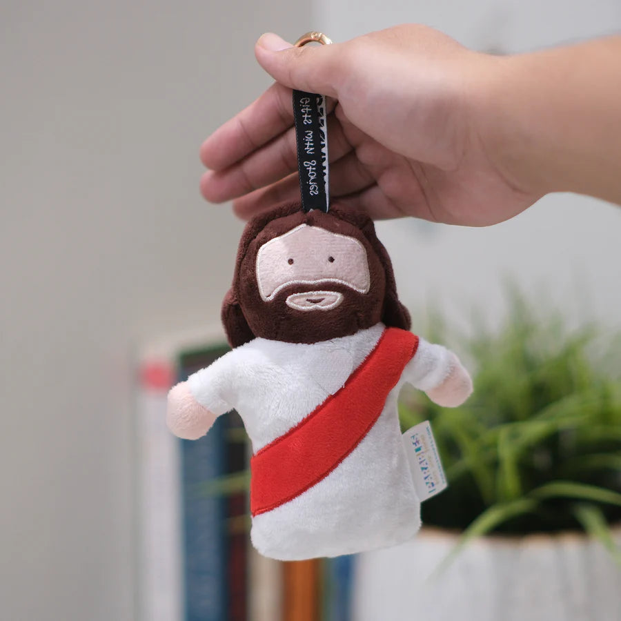 Jesus plushie charm keychain, 6-inch brown plush with white robe and red sash, held by hand—perfect as a faith-based backpack clip and gift.