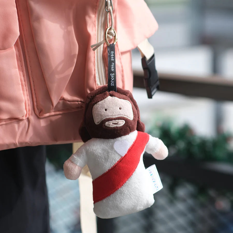 Jesus plushie charm keychain clipped to a pink backpack; soft brown plush with a white robe and red sash, a faith-based backpack charm.