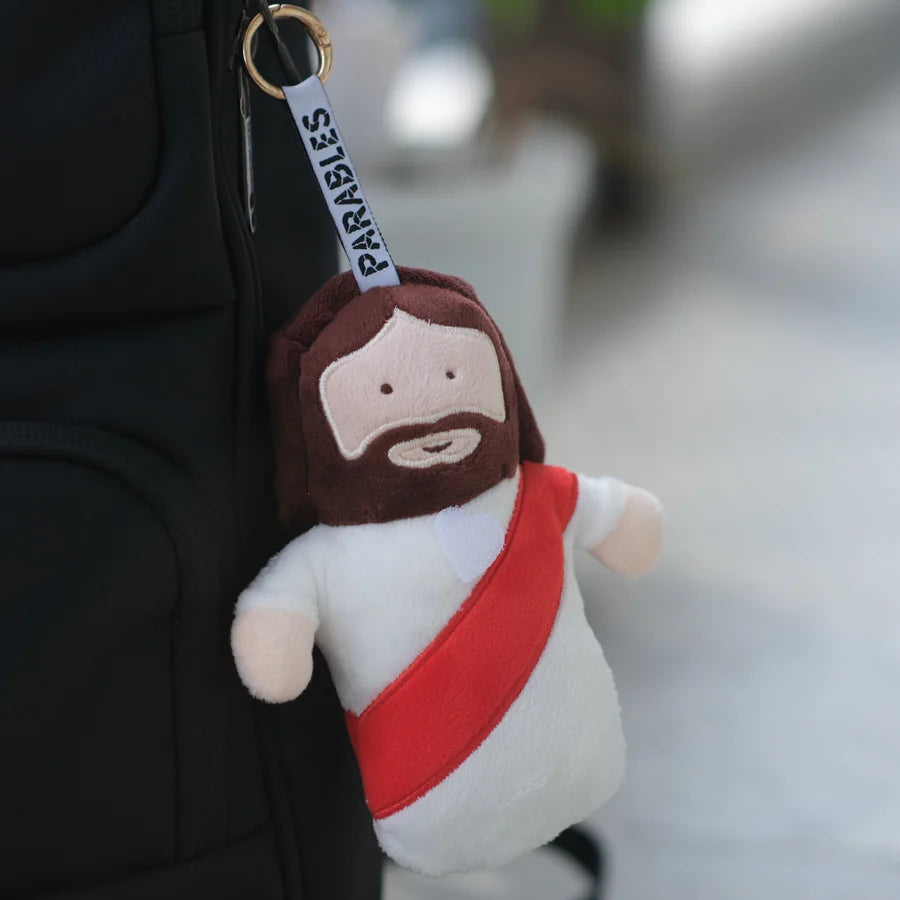 Jesus plushie charm keychain attached to a backpack, soft plush with white robe and red sash
