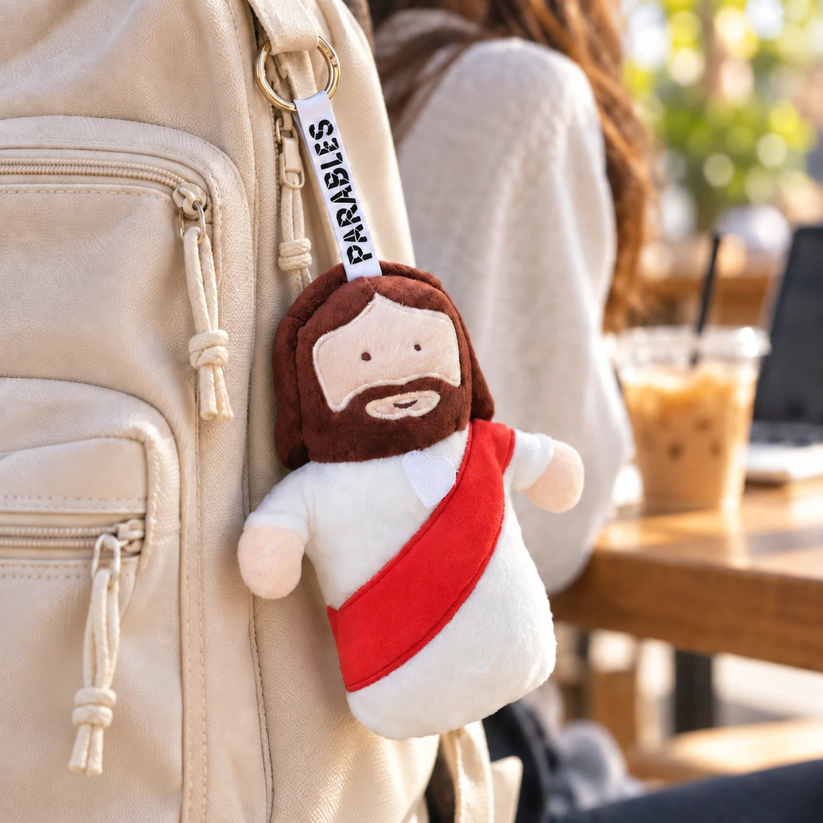 Jesus plushie charm by Parable Gifts clipped to a cream backpack in a warm cafe lifestyle setting.