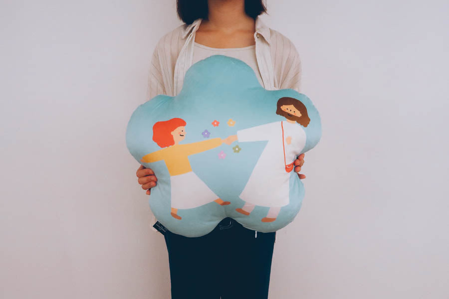 Person holding a 'Joyful Dance with Jesus' plush toy cushion, featuring an illustration of a child dancing with Jesus, symbolizing joy and faith.