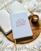 Off-white hard cover journal with “let every glimpse of His grace awaken our hearts in joyous praise” text by Project J placed on wooden tray beside open notebook and candle.