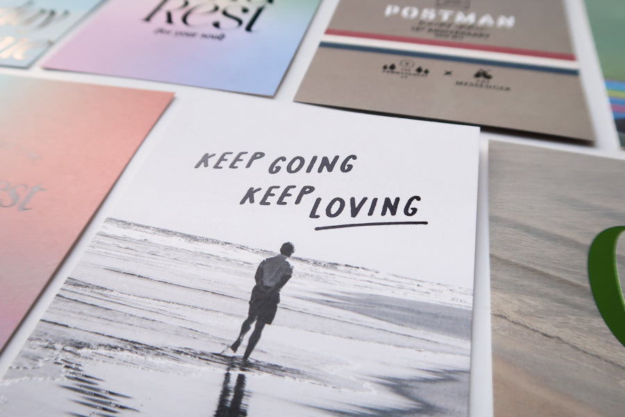 A close-up view of a collection of postcards, with a prominent black-and-white card featuring a person walking on the beach and the motivational message "Keep Going, Keep Loving" along with other faith-based and inspirational designs.