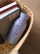 Lavender milk bottle-shaped tumbler with the words “you are so loved” and a small heart, placed in a wicker basket beside books — The Commandment Co.